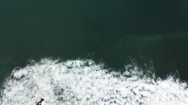 Aerial view of ocean waves crashing against rocks. Surfer rides wave with focused determination. Deep green water contrasts with frothy white foam. Scene captures raw power of nature's movement