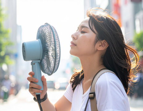 young japanese woman using a portable handheld electric fan outdoors on a hot summer day to cool down