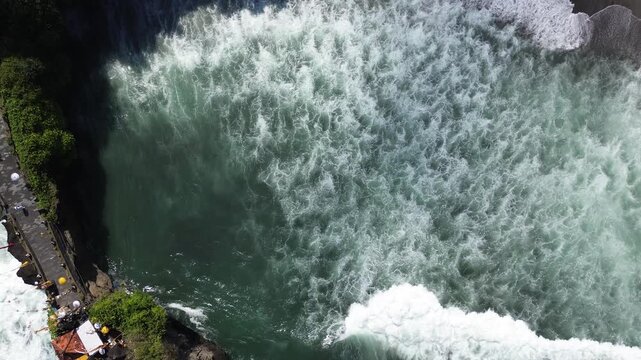 Powerful ocean waves crash against rugged coastline. Vibrant turquoise water churns with frothy white surf. Lush greenery lines the rocky shore. Small structures cling to the cliffside