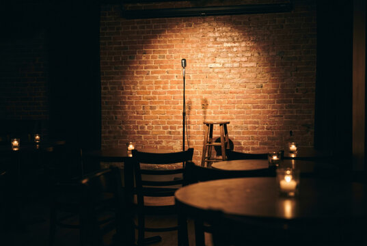 Empty comedy stage with a microphone and stool under a warm spotlight, framed by brick walls, candles and dark club seating