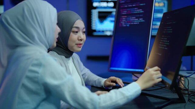 Two young Muslim women in hijabs collaborating on software code in a dark high-tech office. Female programmers analyzing data on dual monitors, smiling during teamwork. Diverse IT professionals.