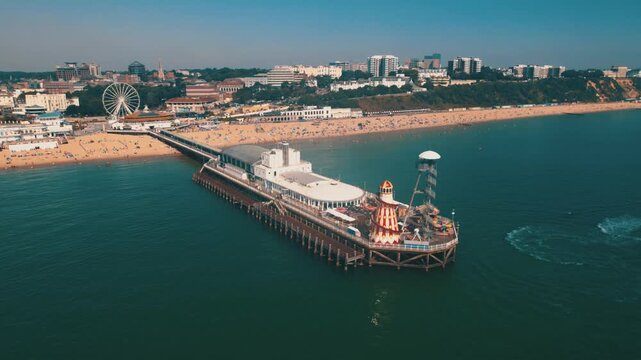Aerial drone shot showing Brighton Palace Pier with amusement rides, busy sandy beach, seafront buildings and calm blue water along Brighton coast in England