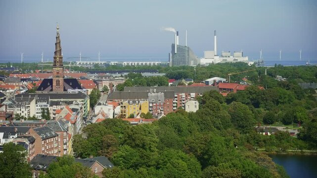 Aerial panorama of Copenhagen featuring the historic Church of Our Saviour, modern Amager Bakke power plant, and offshore wind turbines blending green energy with urban heritage.
