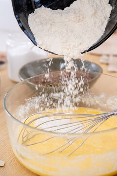 Dough making. Pouring flour. Adding ingredient to yellow batter in glass bowl with whisk on wooden table. Baking culinary process.