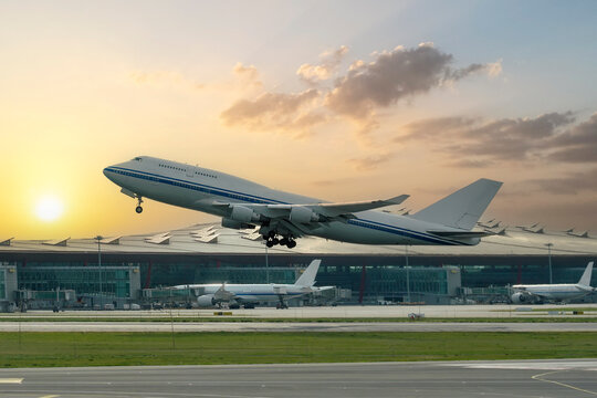 View of a commercial airline passenger airplane taking off from the runway, with the airport terminal building structures, against the backdrop of the rising sun and clouds