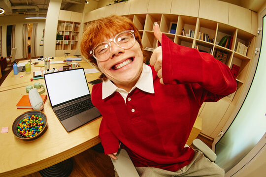 Portrait of young adult Caucasian man with glasses showing thumbs up gesture at office desk with laptop. Image represents positive feedback and remote work success, wide angle shot
