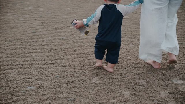 Low level shot of toddler taking steps with mother walking along ocean shore.
