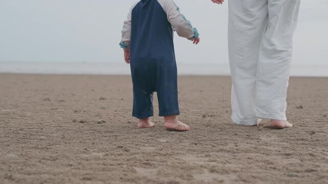 Asian mother holding hands with toddler son walking on sandy beach together.