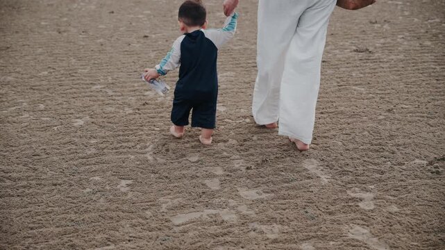 Asian mother holding hands with toddler son walking on sandy beach together.