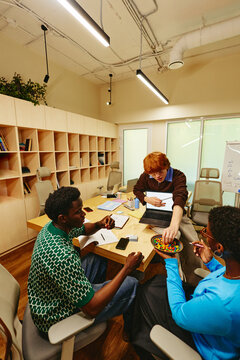 Vertical image of diverse young adult colleagues collaborating around office table with laptop and sharing snacks, perfect for corporate diversity and modern workplace culture campaigns