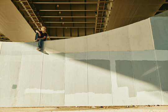 Caucasian Gen Z man sitting on concrete wall under bridge, representing youth culture and urban lifestyle. Ideal for youth-focused marketing campaigns, wide angle shot