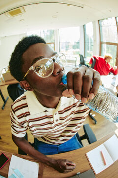 Young adult African American man wearing glasses drinking water from plastic bottle in office. Image suitable for healthy lifestyle and workplace wellness campaigns, vertical