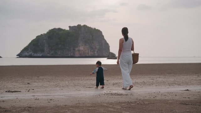 Asian mother holding hands with toddler son walking on sandy beach together.