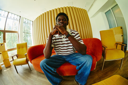 Portrait of young adult African American man sitting on sofa in modern office lobby, looking at camera. Image represents corporate diversity and recruitment, wide angle shot