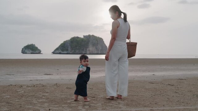 Asian mother holding hands with toddler son walking on sandy beach together.