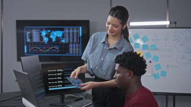 Asian tech lead points at a data dashboard while holding a green circuit board to explain hardware integration to a seated developer in a modern technology office.