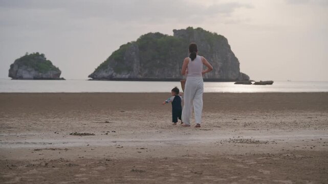 Asian mother holding hands with toddler son walking on sandy beach together.