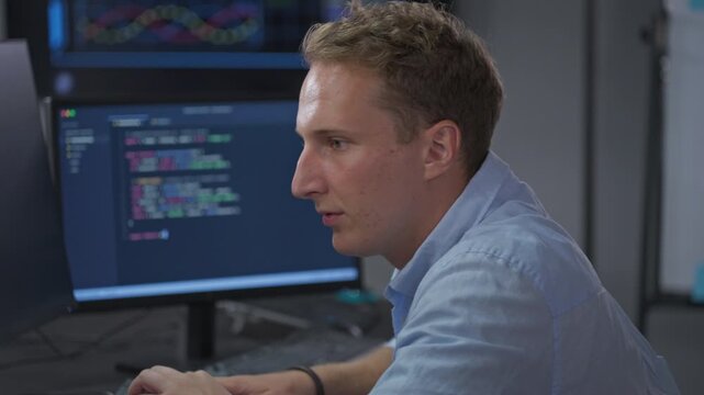 Close side view of a young male developer studying lines of code on his monitor with total concentration during a focused software development session in a dim office.