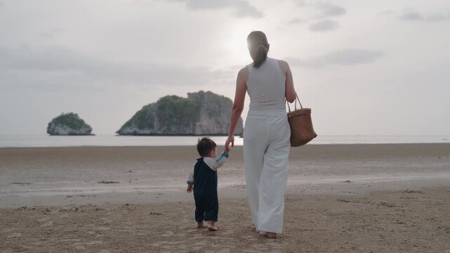 Asian mother holding hands with toddler son walking on sandy beach together.