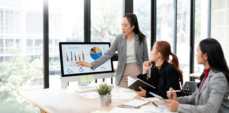 Asian Business Woman Presenting Financial Charts on Computer Screen to Team in Office