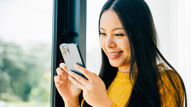 Young woman smiling uses a smartphone app by the window typing and connecting for social media at home. Reflecting communication online reading and texting.