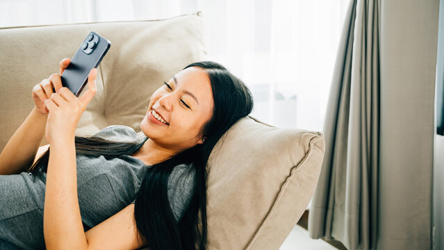 Asian woman on couch texts on smartphone smiling. Enjoying relaxation chatting and online shopping. Modern technology for communication and connection at home.