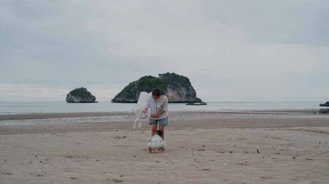  Asian mother holding hands with toddler son standing on beach.