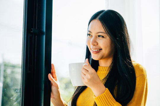Morning delight, Smiling woman stands by the window holding a cup of coffee enjoying sunrise at home. Reflecting relaxation happiness and contemplation at dawn.