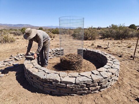 Gardener builds circular stone garden bed with wire mesh compost bin in arid landscape