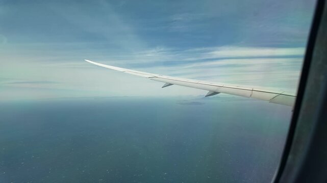 Airplane wing flies over blue ocean view from cabin window.