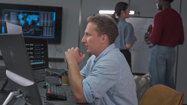 Young male developer rests his chin on his hand while thinking through data dashboards on his monitors, with teammates working at a whiteboard behind him in the office.