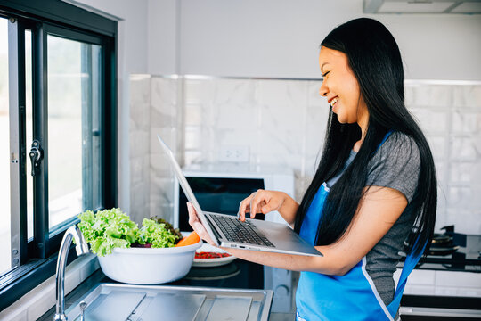 In her kitchen a smiling woman enjoys cooking while exploring recipes on her laptop. Portraying the joy and convenience of technology in meal preparation.