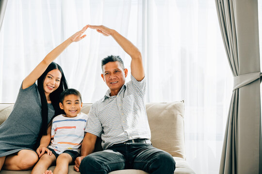 A smiling family of four on a sofa parents making roof gestures over their little son. Representing home security children protection and future plans ensuring family safety.