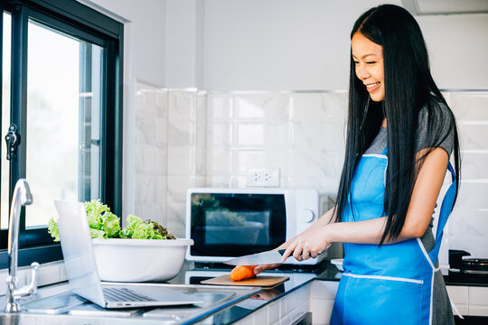 In the kitchen a smiling woman stands engrossed in cooking and using her laptop. Portraying the joy of technology in culinary experiments and meal preparation.