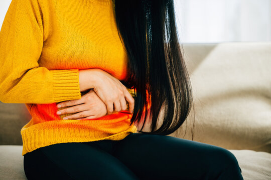 An Asian woman in living room at home endures stomach ache on a couch. Reflecting discomfort symptoms and the importance of medical care for stomachache. Health care abdomen bloating concept