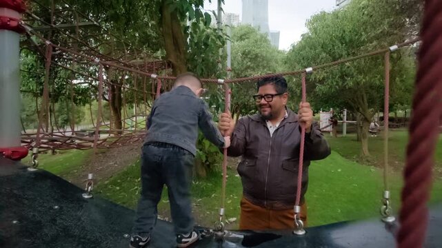 A happy Mexican father in a leather jacket watches his two-year-old son in a denim outfit jump and laugh on a rope bridge at a children's playground. The video features a zoom-in shot, capturing the j