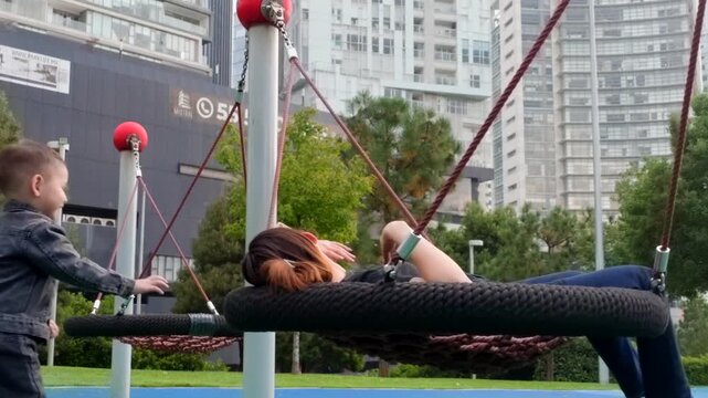 A joyful scene unfolds as a two-year-old boy playfully pushes his young Latin mother on a modern swing bed at a vibrant city park playground. Their interaction is filled with laughter and warm embrace