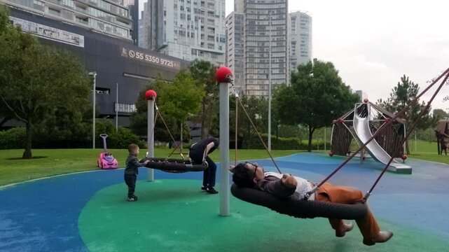 A joyful 2-year-old toddler enjoys playing on modern swing beds with his young Latina mother and dark-haired father at an urban park playground. The family shares laughter and hugs, cherishing their q
