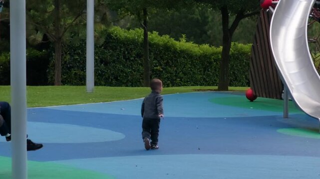 A cute two-year-old boy, dressed in warm clothes, waits patiently for his mother at the entrance of a vibrant playground on a chilly day, eager to play.