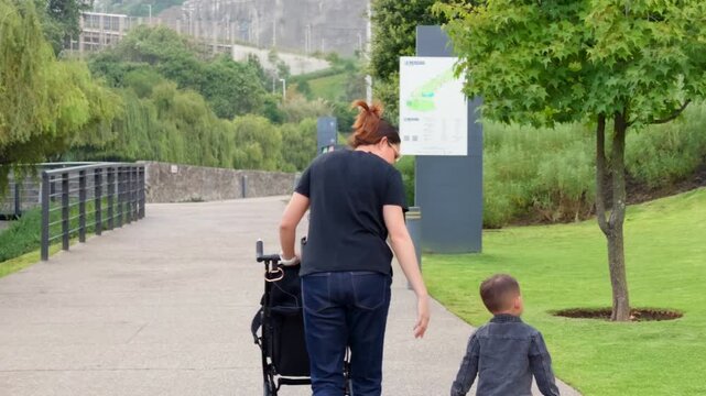 A young Latina mother and her 2-year-old son walk from behind with a stroller on a paved path in a lush green park, enjoying the serene natural environment on a pleasant day.