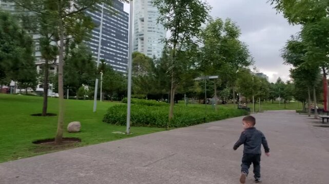A young Mexican-Latina couple and their 2-year-old baby, dressed in a jumpsuit, run cheerfully along a concrete path in a vibrant urban park. The baby happily looks at its parents, who are pushing a s