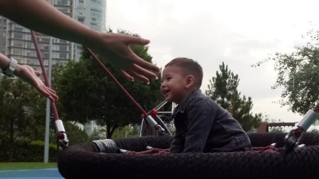 A close-up of a young Latina mother tenderly pushing her smiling 2-year-old baby on a swing bed at a vibrant park playground, filled with genuine laughter and affectionate hugs.