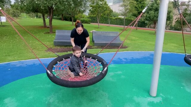 A young Latina mother gently pushes her happy 2-year-old toddler on a large nest swing in a vibrant public park playground. The scene is filled with laughter and affectionate hugs, highlighting a joyf