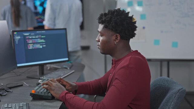 Young Black software developer types code at his workstation while colleagues move in the background, with an agile sticky note board on the wall of a modern tech office.