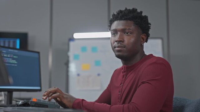 Young Black developer types at his keyboard while looking toward the camera, with code on his monitor and a sticky note agile board on the wall of a modern tech office.