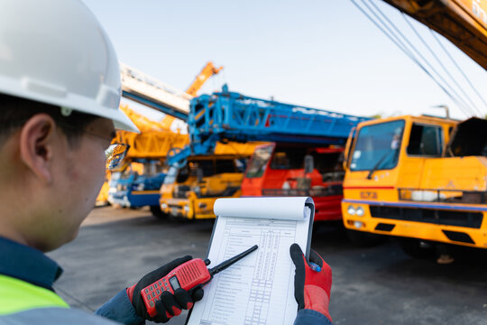 In a crane yard, an inspection of heavy machinery is conducted by a male engineer holding his radio and clipboard