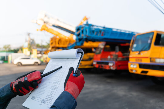 At a lifting operations site, a safety inspector wearing protective gloves holds a radio and clipboard to review heavy machinery