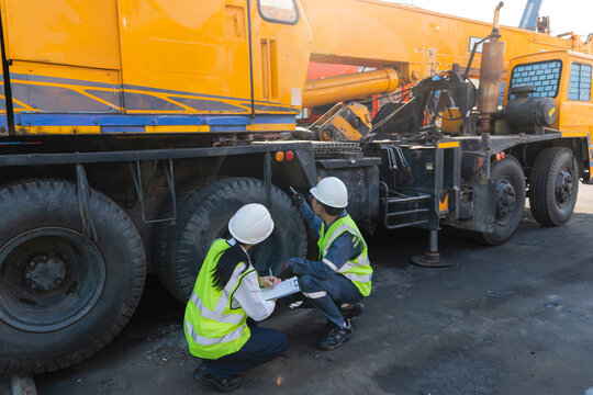 At the lifting equipment inspection site, two workers wearing hard hats squat to check yellow crane tires using one clipboard