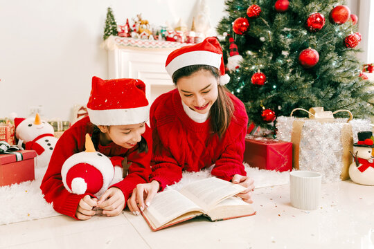Happy mother and young daughter wearing Santa hats and reading a book together beside a decorated Christmas tree, sharing a cozy, loving moment during the joyful festive holiday season.