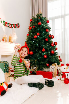 Smiling child in an elf hat sits by a decorated Christmas tree, holding milk and a cookie, surrounded by gifts and festive decor in a cozy living room.